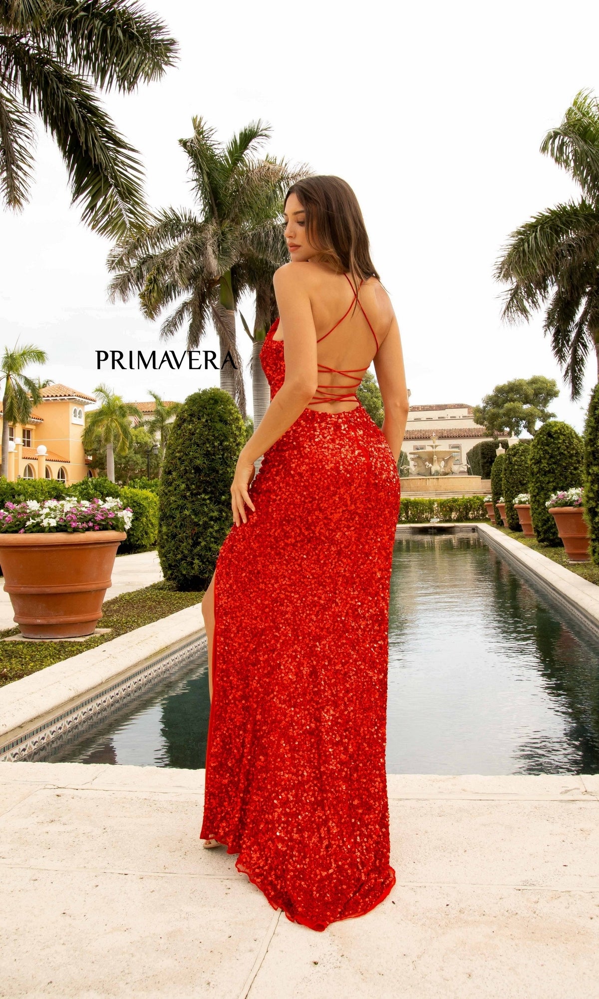 A woman in an Open Back Bold Long Sequin Prom Dress 3290D stands with her back to the camera by a pool, surrounded by palm trees and manicured hedges.